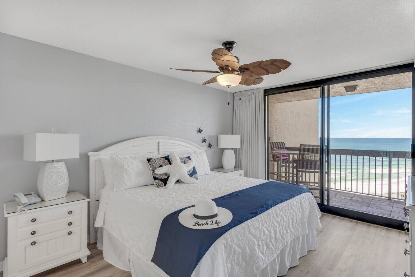 Ein Hotelzimmer mit einem Bett, einem Deckenventilator, einem Nachttisch mit Lampe sowie einem weiteren Nachttisch mit Telefon und Blick auf den Strand durch ein großes Fenster.