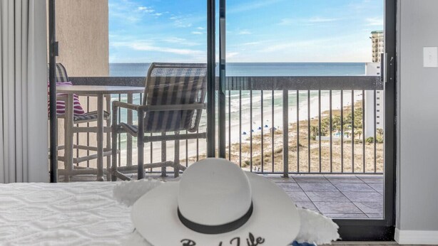 A bedroom with a bed, a white hat with 'Beach Life' text, a view of the beach and ocean through a balcony door. - Spectacular views of the Gulf and Emerald coast - 16th floor. (Destin)
