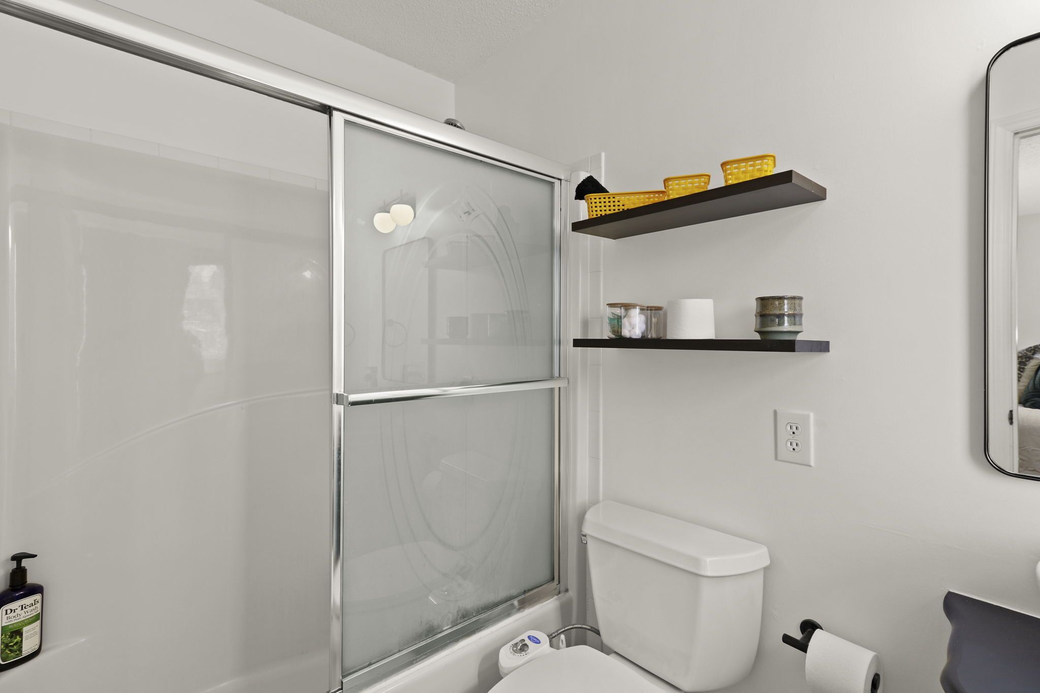 Ein modernes Badezimmer mit Duschabtrennung aus Glas, Toilette, Ablage für Pflegeprodukte und Spiegel an der Wand.