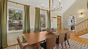 Un comedor con una mesa y sillas de madera, una araña y vistas al exterior a través de amplias ventanas.