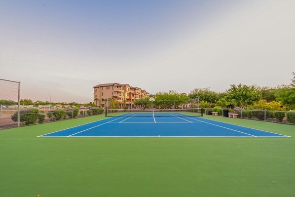 An outdoor tennis court with a net, surrounded by greenery and a building in the background.