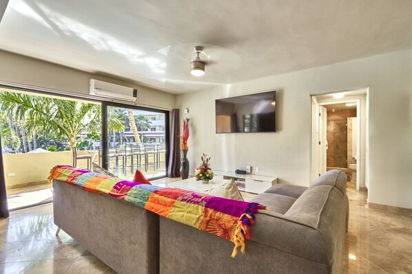 A modern living room with a sofa, a flat-screen TV, and a view of palm trees through large windows. - Three Bedroom Condo Pool View, one block away from Medano Beach (Cabo San Lucas)