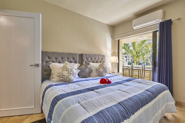 A bedroom with a large bed, a grey tufted headboard, and a view of the outdoors through a window with curtains. - Three Bedroom Condo Pool View, one block away from Medano Beach (Cabo San Lucas)