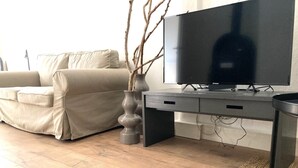 Olohuoneessa on beigenvärinen sohva, harmaalla jalustalla oleva taulutelevisio ja maljakko, jossa on oksia.
