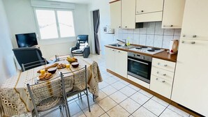 A modern kitchen with white cabinets, a dining table with a patterned tablecloth, and a TV in the background. - Ronsard (Bains-les-Bains)