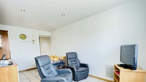 A room with two reclining chairs, a small round table, a television on a stand, a wooden cabinet, and a tiled floor. - Ronsard (Bains-les-Bains)