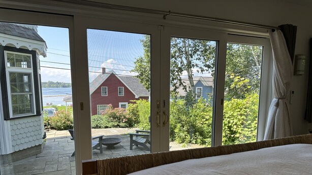 Et soveværelse med en stor seng, en skydedør til en terrasse med udendørs møbler, samt udsigt til et rødt hus og grøn vegetation.