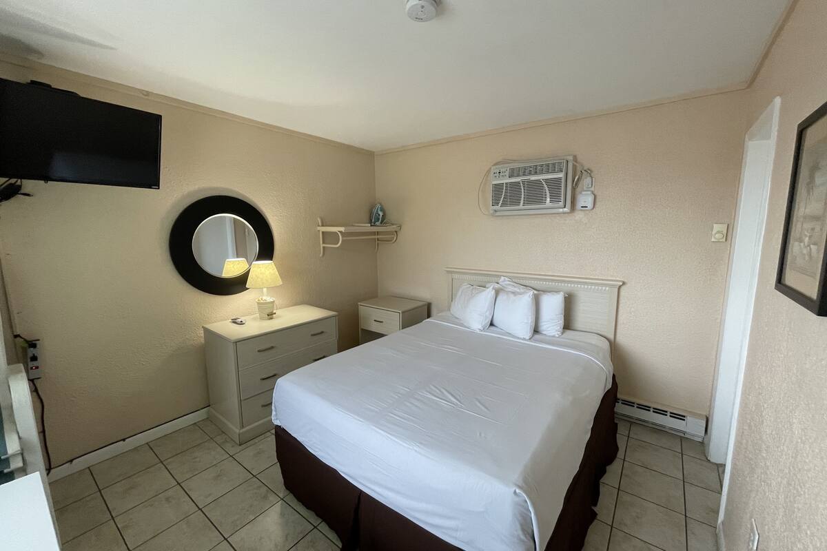 A hotel room with a bed, a bedside table, a mirror, a wall-mounted air conditioner, and a ceiling light.