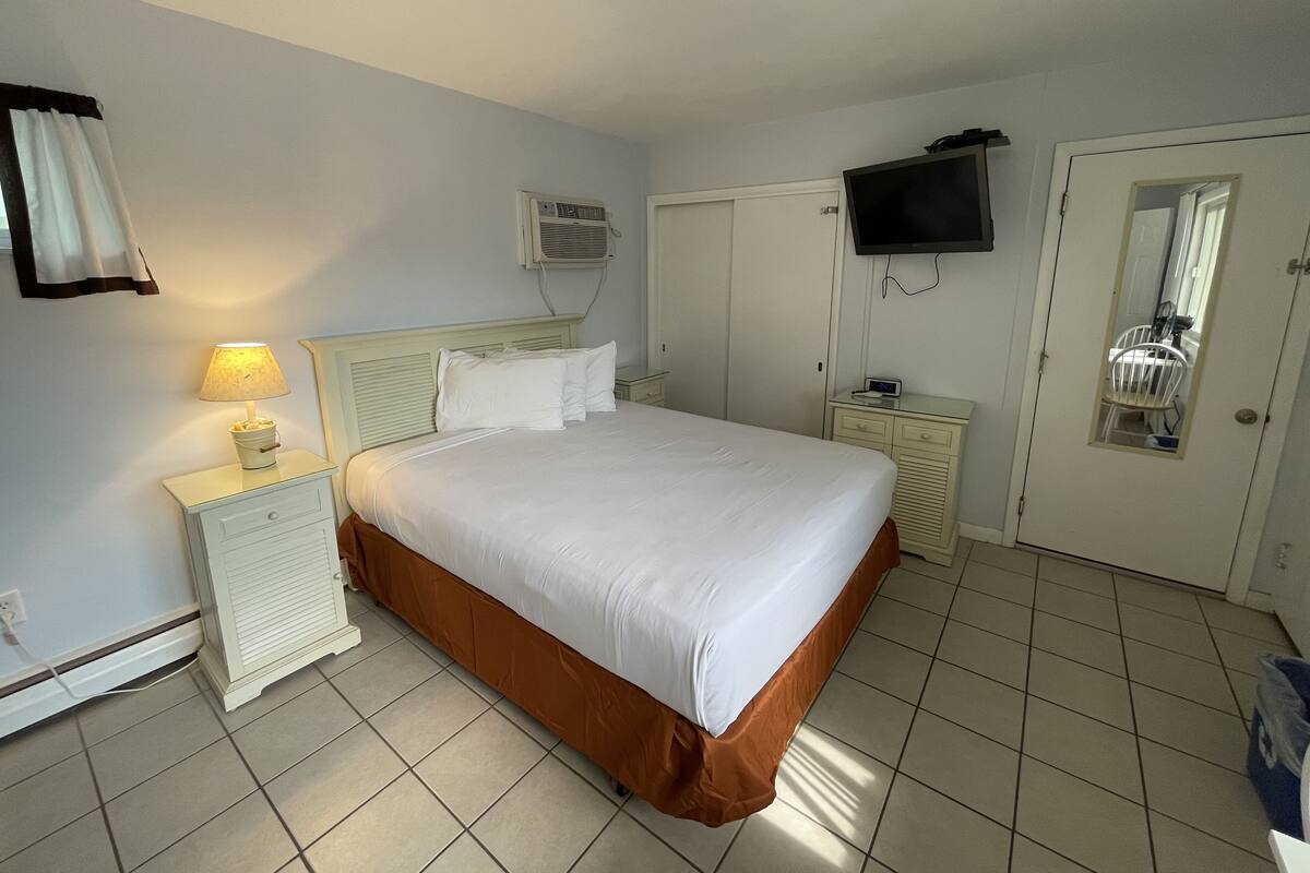 A hotel room with a bed, a bedside lamp, a TV, a mirror, and a door with a chair inside.