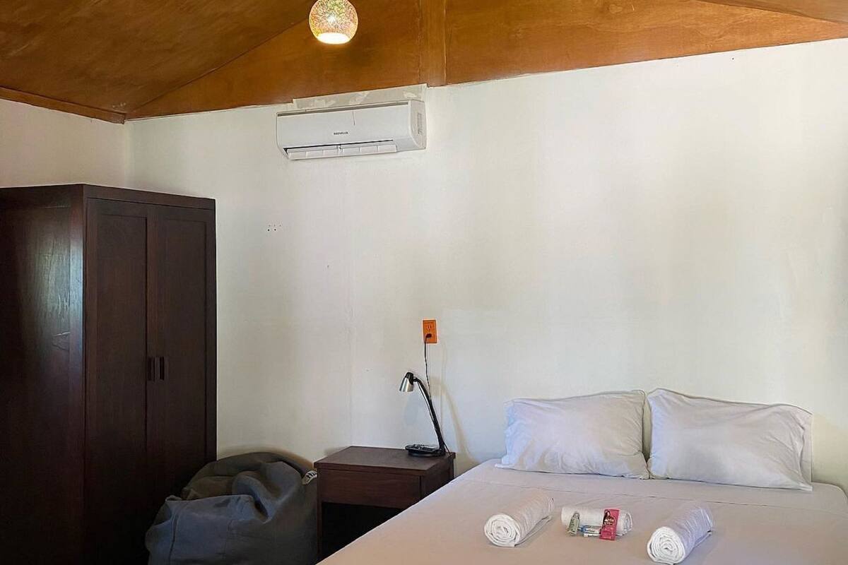 Une chambre avec un plafond en bois, un lit recouvert de draps blancs, une lampe de chevet, une armoire et un appareil de climatisation.