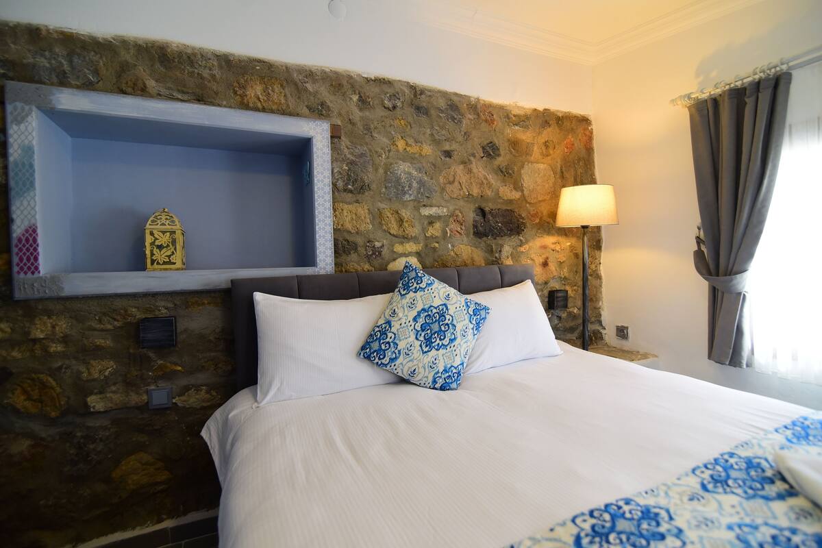 A hotel room with a stone wall, a bed with white bedding and blue patterned pillows, a nightstand with a lamp, and a small shelf with a decorative item.