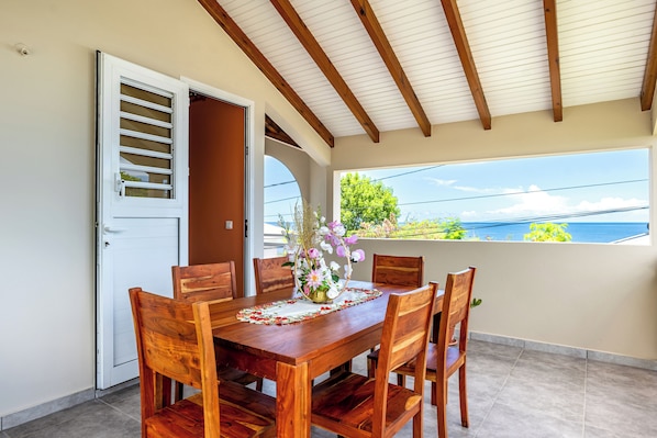 A wooden dining table with six chairs, a vase with flowers, and a view of the sea through a window.