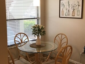 A dining area with a glass table surrounded by wicker chairs, a vase with a plant, and a framed artwork of vases on the wall.
