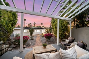 A covered patio area with outdoor furniture, a pathway leading to a house, and a garden with various plants and a cactus.