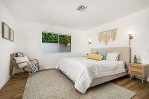 A bedroom with a bed, a chair, a nightstand, a wall-mounted lamp, and a window with a view of greenery. - Newly Renovated Palm Springs Oasis (Palm Springs)