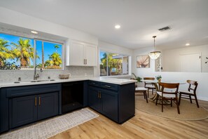 A modern kitchen with dark cabinetry, a large island, and a dining area with wooden chairs and a glass table. - Newly Renovated Palm Springs Oasis (Palm Springs)