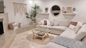 Un salon moderne avec un canapé beige, une table basse en verre et une cheminée. On y trouve des plantes décoratives, une décoration murale en macramé et un miroir rond accroché au mur.