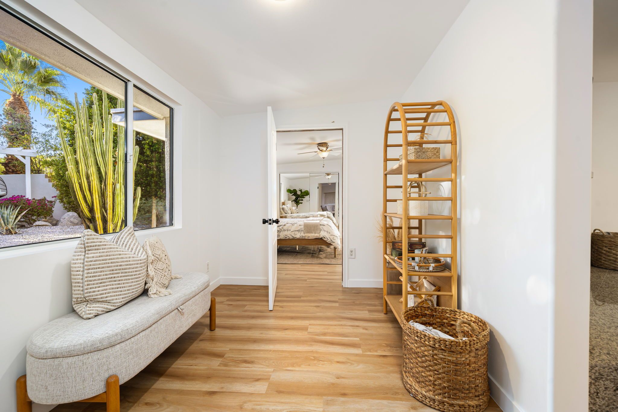 A hallway with a wooden floor, a white wall, a window with a view of cacti, a white bench with cushions, and a wooden ladder shelf.