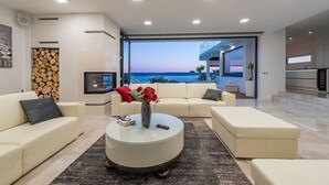 A modern living room with a large window overlooking the sea, a fireplace, and a round coffee table with a vase of flowers.