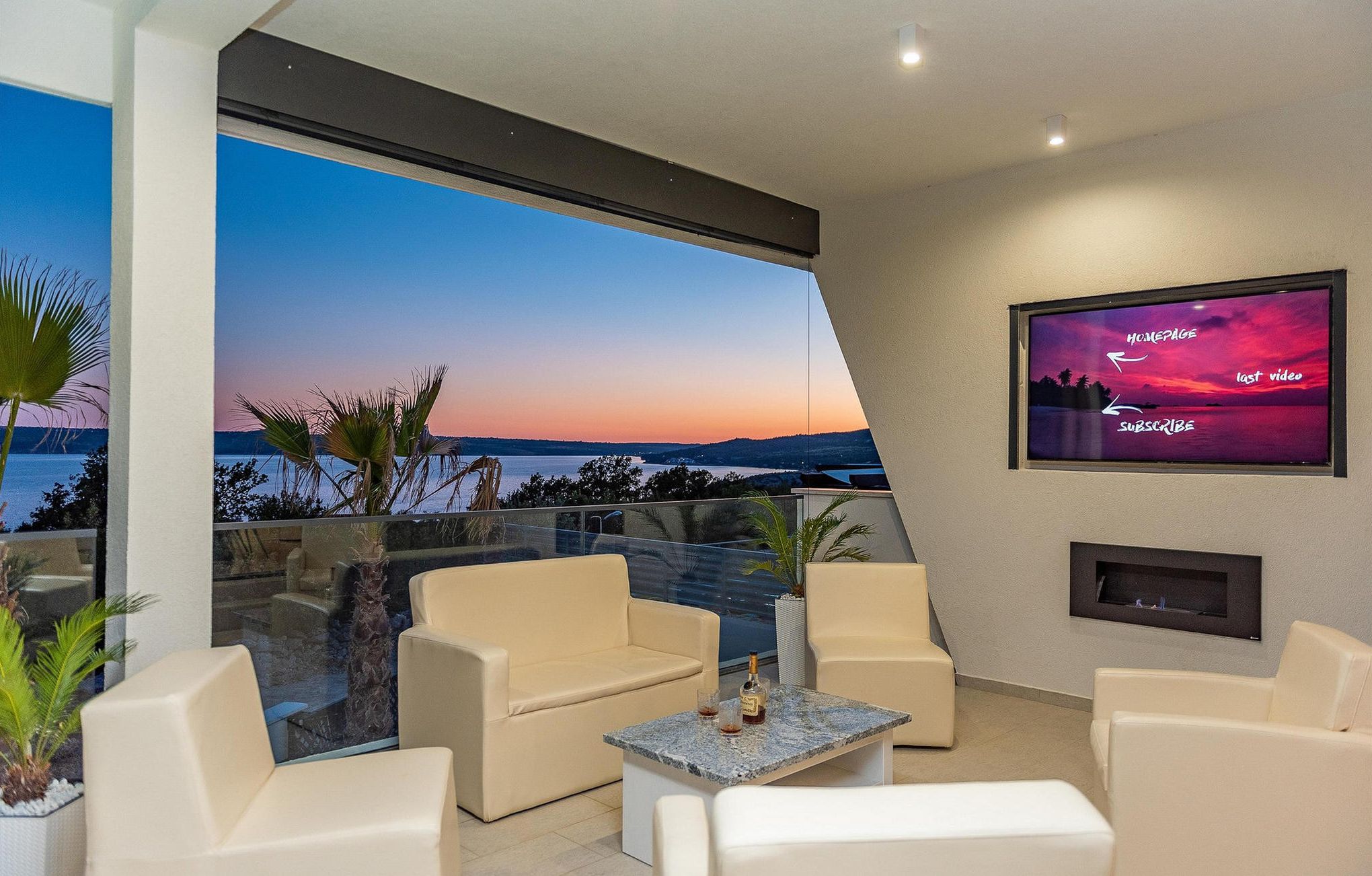 A modern living room with a large window overlooking a scenic view, comfortable seating, and a mounted TV displaying a sunset scene.