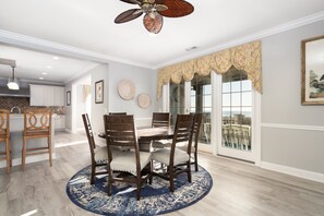 A dining area with a round table, six wooden chairs, and a blue patterned rug. - Footprints On The Sand: Private Beach, Pools & Tiki Bar (Murrells Inlet)