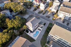 An aerial view of a residential area with a large house featuring a swimming pool, a driveway, and a street with parked cars.
