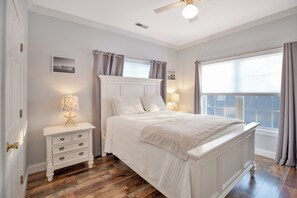A bedroom with a large bed, a nightstand with a lamp, a ceiling fan, and a window with curtains.