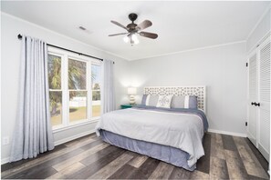 Un dormitorio con cama, ventilador de techo, ventana con cortinas y mesita de noche con lámpara.