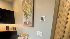 Et kompakt køkken med kaffemaskine, brødrister og en lille ø med en vase med blomster. Der er et maleri af solsikker på væggen, en termostat og en dør til højre.