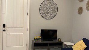 En stue med en hvid dør, et gråt tv-møbel med et fladskærms TV, en blå sofa med en gul pude og en rund dekorativ vægdekoration.