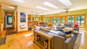 A spacious living area with a large sofa, a coffee table, and a dining table. There is a kitchen area with cabinets and a window overlooking a scenic outdoor view. - Waterfront Home -  Mid Coast Maine near Camden and Belfast (Belmont)