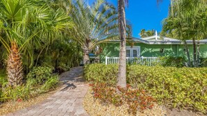 A pathway leading to a green building with a white fence, surrounded by palm trees and various shrubs. - Key Lime Cottage by Beach Boutique Rentals (Holmes Beach)