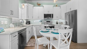 A modern kitchen with white cabinets, a stainless steel refrigerator, a microwave, and a dishwasher. There is a dining table with chairs and a small island with a sink. - Key Lime Cottage by Beach Boutique Rentals (Holmes Beach)