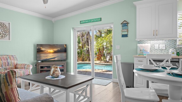 A living room with a TV, a dining table set for two, a sofa, and a view of a pool and palm trees through the glass door. - Key Lime Cottage by Beach Boutique Rentals (Holmes Beach)