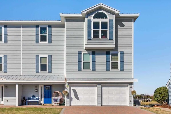 A two-story residential building with a garage, a porch with seating, and a small garden area. - The Blue Heron Hideaway (Tybee Island)