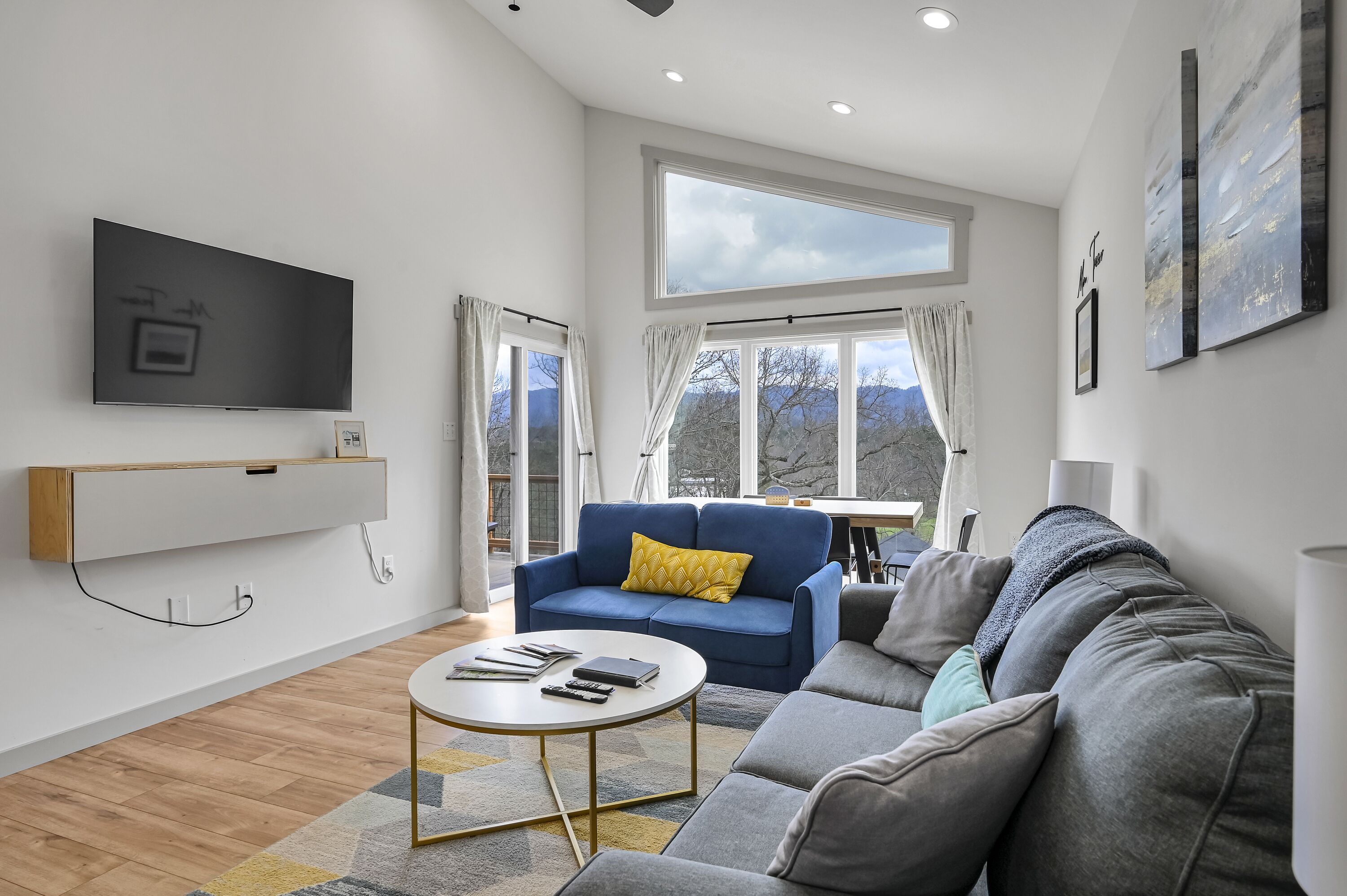 A modern living room with a flat-screen TV, a sofa, a coffee table, and a large window with a view.