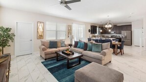 A modern living room with a beige sofa, a coffee table, and a blue rug. There is a ceiling fan, a dining area with a chandelier, and a kitchen with dark cabinets.