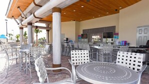 An outdoor patio area with metal chairs and tables, a bar area with a TV, and a pool in the background.