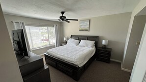 A bedroom with a bed, bedside tables, a ceiling fan, a window with blinds, and a TV on a stand.