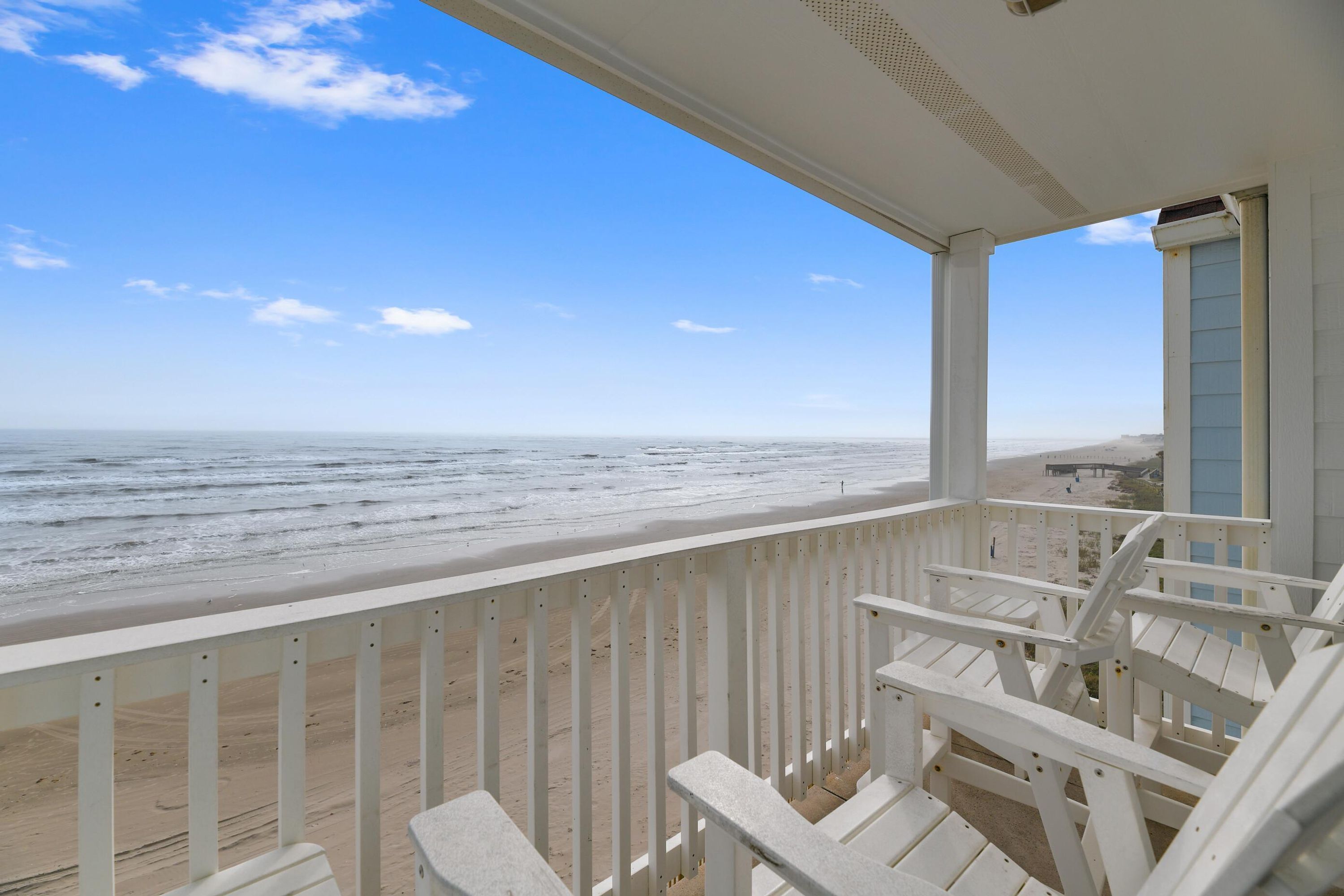 A beach view from a porch with white railings and chairs.
