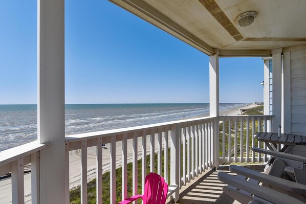 A coastal view from a porch with a white railing, overlooking the beach and ocean.