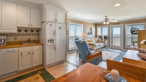 A compact kitchen with white cabinets, a refrigerator, and a coffee maker. There is a dining area with a table, chairs, and a wicker chair. The room has a ceiling fan and a view of the ocean through large windows.