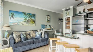 Una sala de estar con un sofá azul, una mesa de centro y un televisor. Cuenta con una cocina equipada con refrigerador, microondas y una encimera de madera.