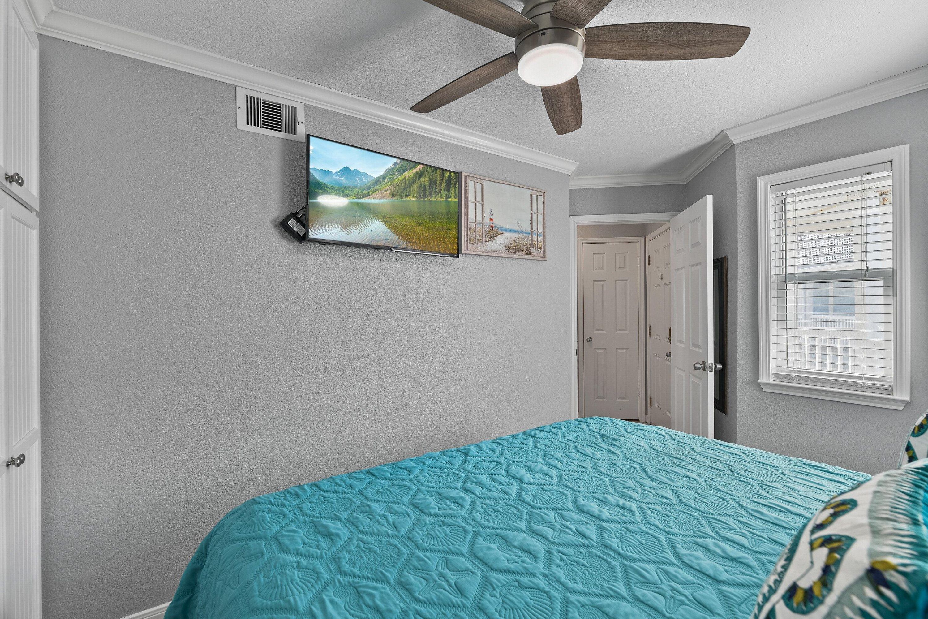 A bedroom with a teal bedspread, a ceiling fan, a TV mounted on the wall, and a window with blinds.