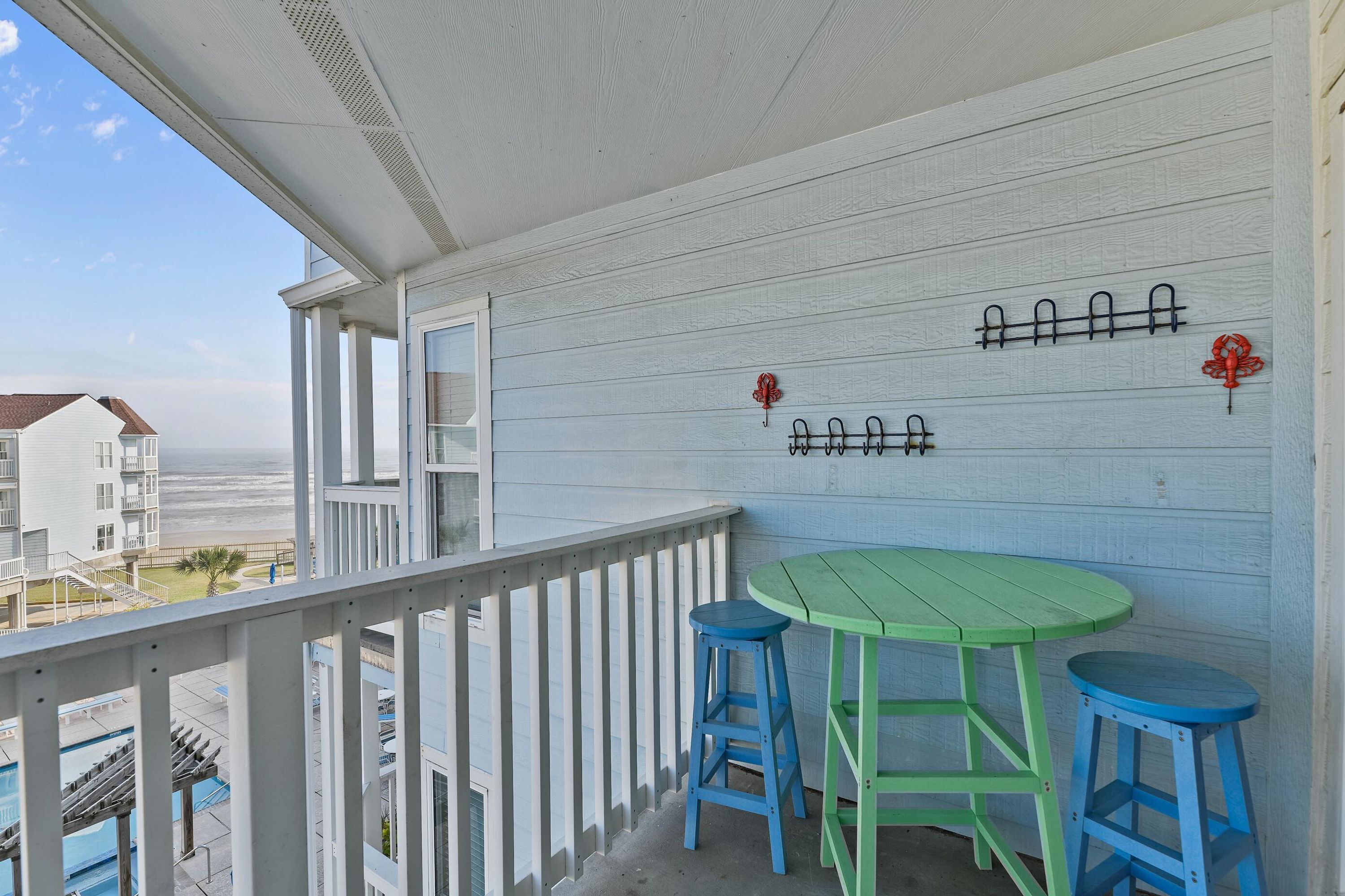 Ein Balkon mit einem grünen Tisch und blauen Hocker, mit Blick auf einen Strand und das Meer.