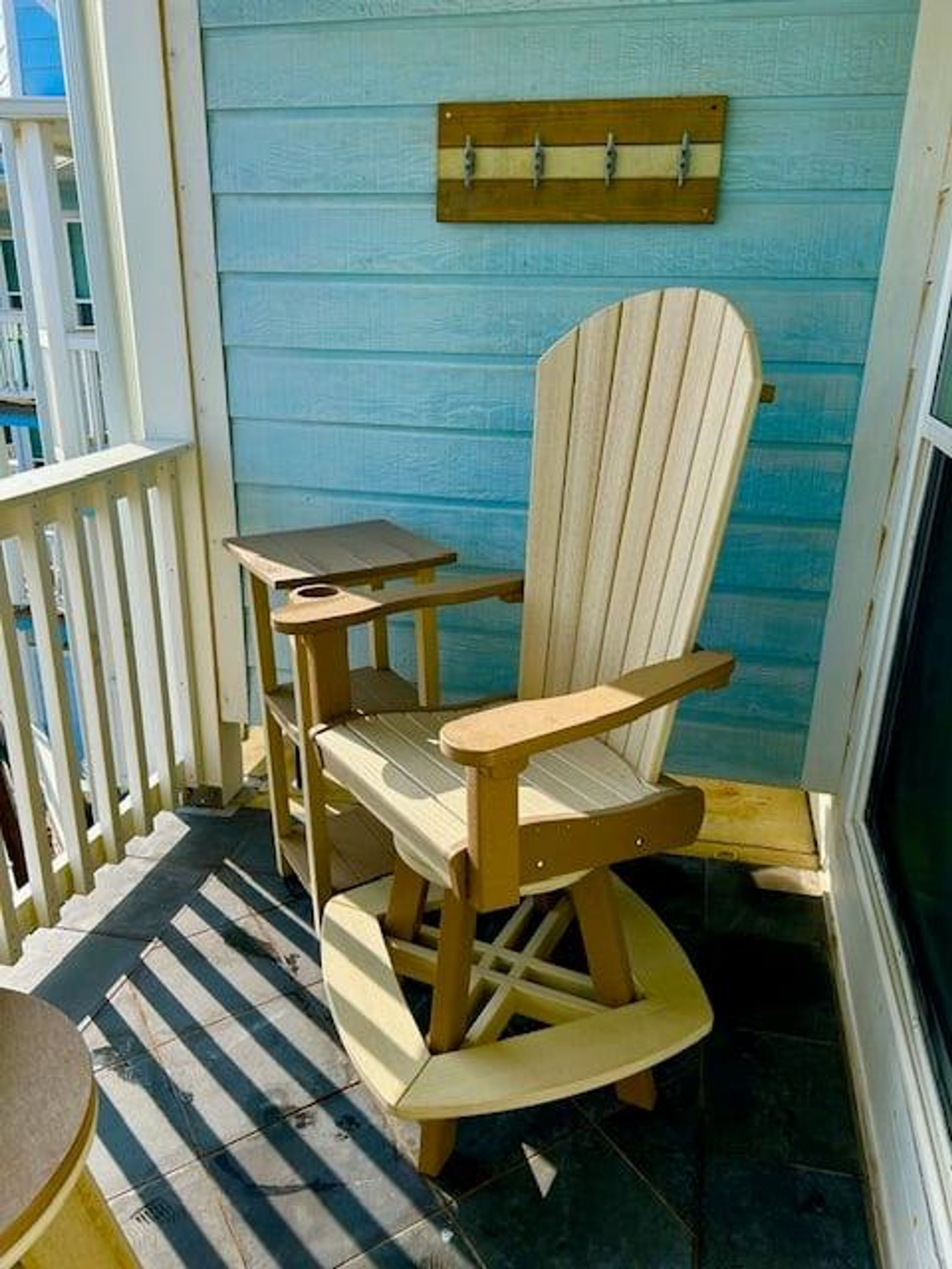 A wooden Adirondack chair with a small table beside it, on a porch with a blue wall and a wooden railing.