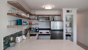 A modern kitchen with white countertops, a stainless steel refrigerator, a microwave, and a stove. There are wooden shelves above the counter holding various kitchen items. - Seascape 2307 in Galveston (Galveston)