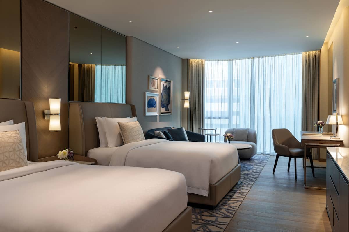 A modern hotel room with two beds, a desk, a chair, and a large window with curtains.