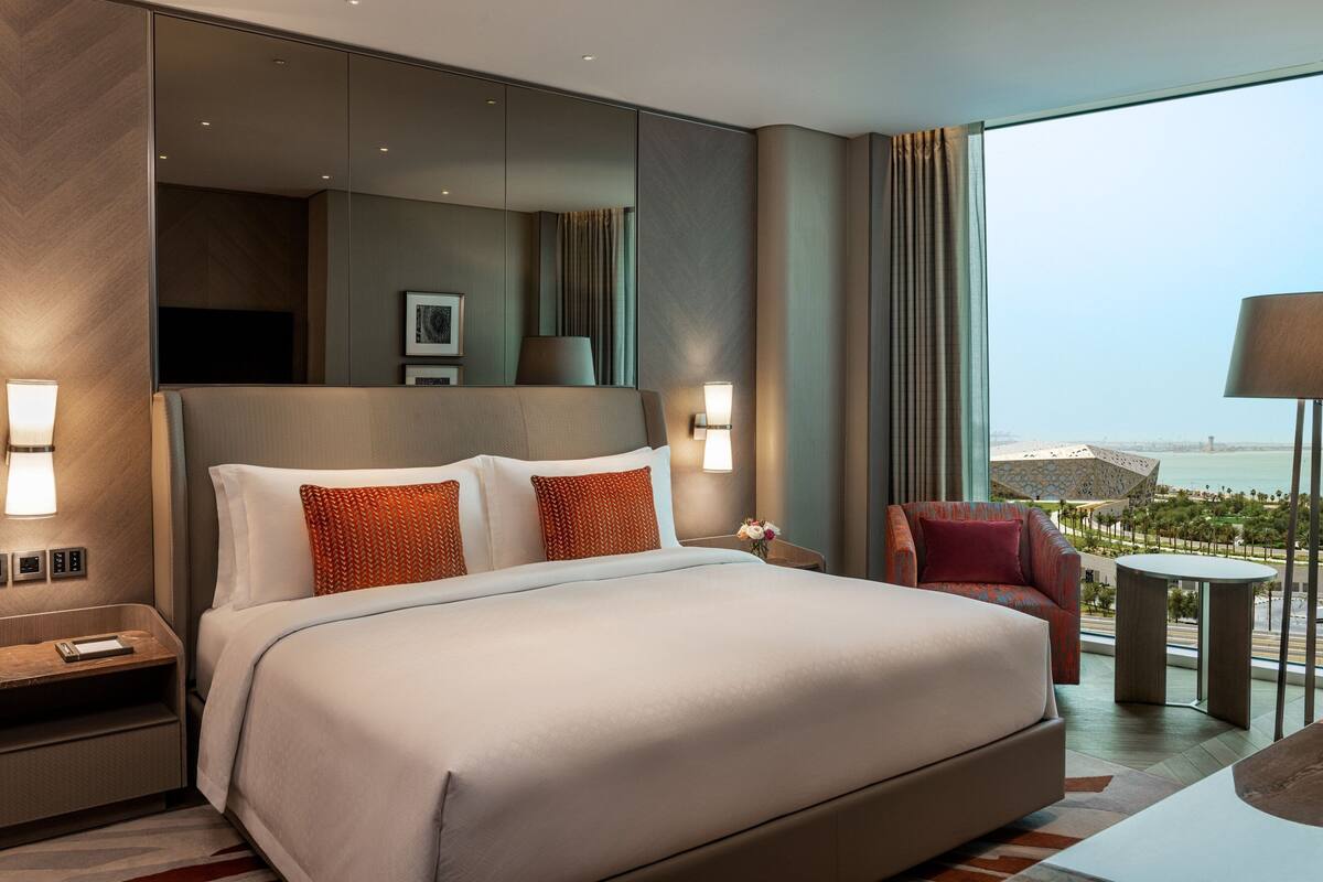 A modern hotel room with a large bed, a sitting area with a chair, a small table, and a large window offering a view of the city.