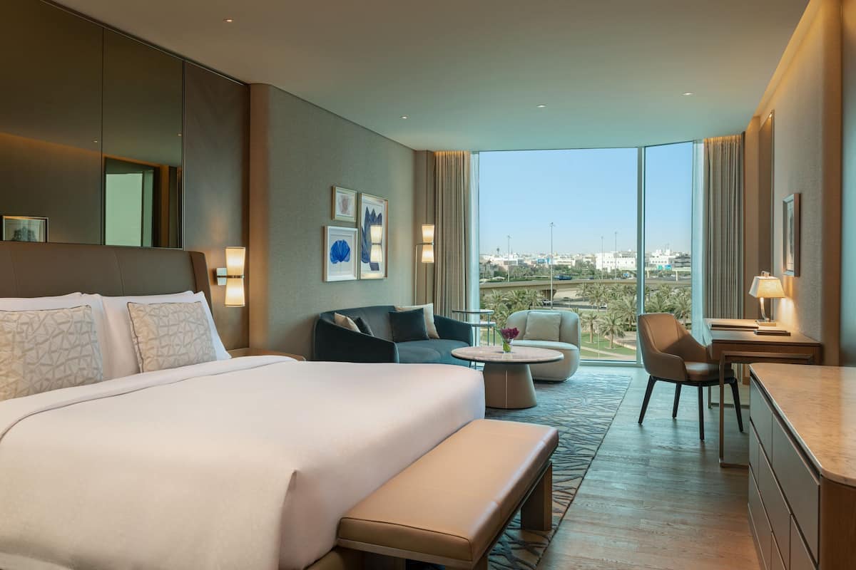A modern hotel room with a large bed, a sofa, a dining area, and a view of the city.